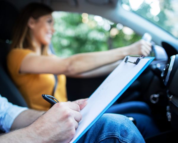 close-up-view-driving-instructor-holding-checklist-while-background-female-student-steering-driving-car